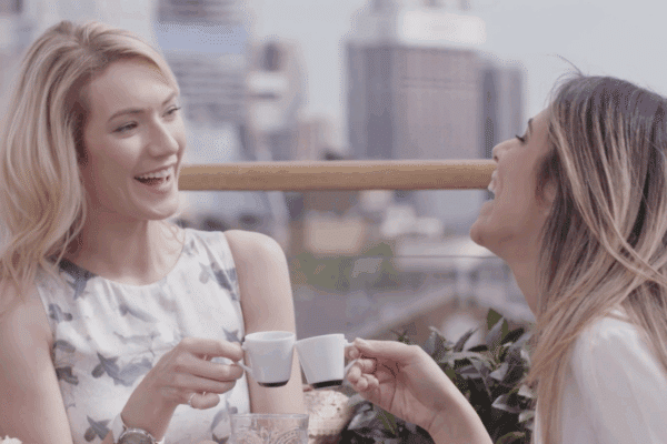 Two women laughing while clinking two small cups with espresso