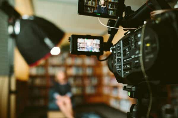an image of a camera in the foreground, with a blurred out light, and person set up for an interview in front of a book case in the background