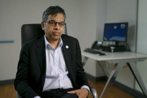 A man of indian heritage in a suit sits in a chair in an interview situation. A computer desk is behind him